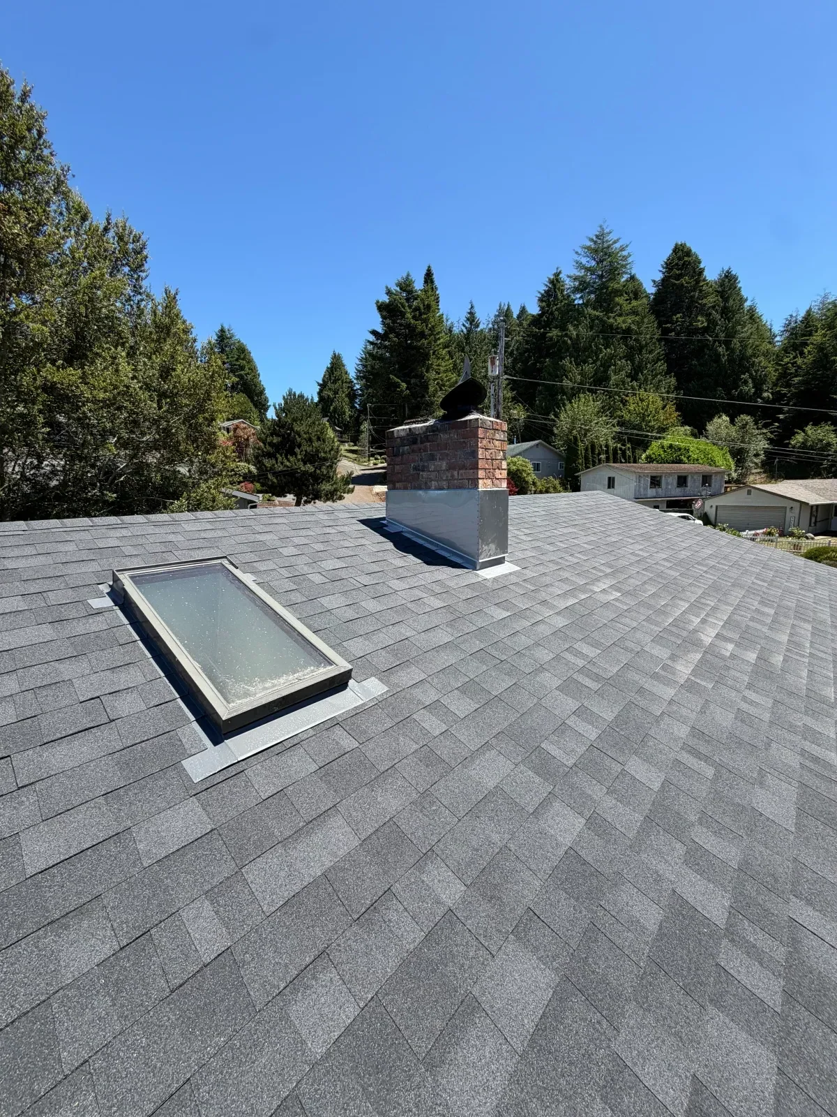 Gray architectural asphalt shingle roof with skylight, metal flashing, and brick chimney with metal chase cover