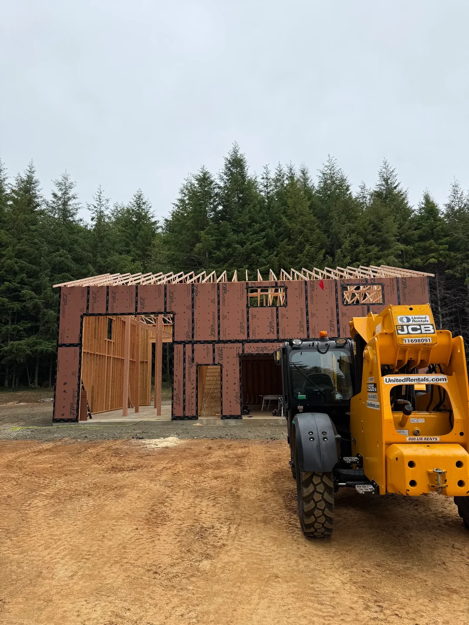 Wood-framed house under construction with ZIP System sheathing, exposed studs, roof trusses, and a JCB telehandler on site