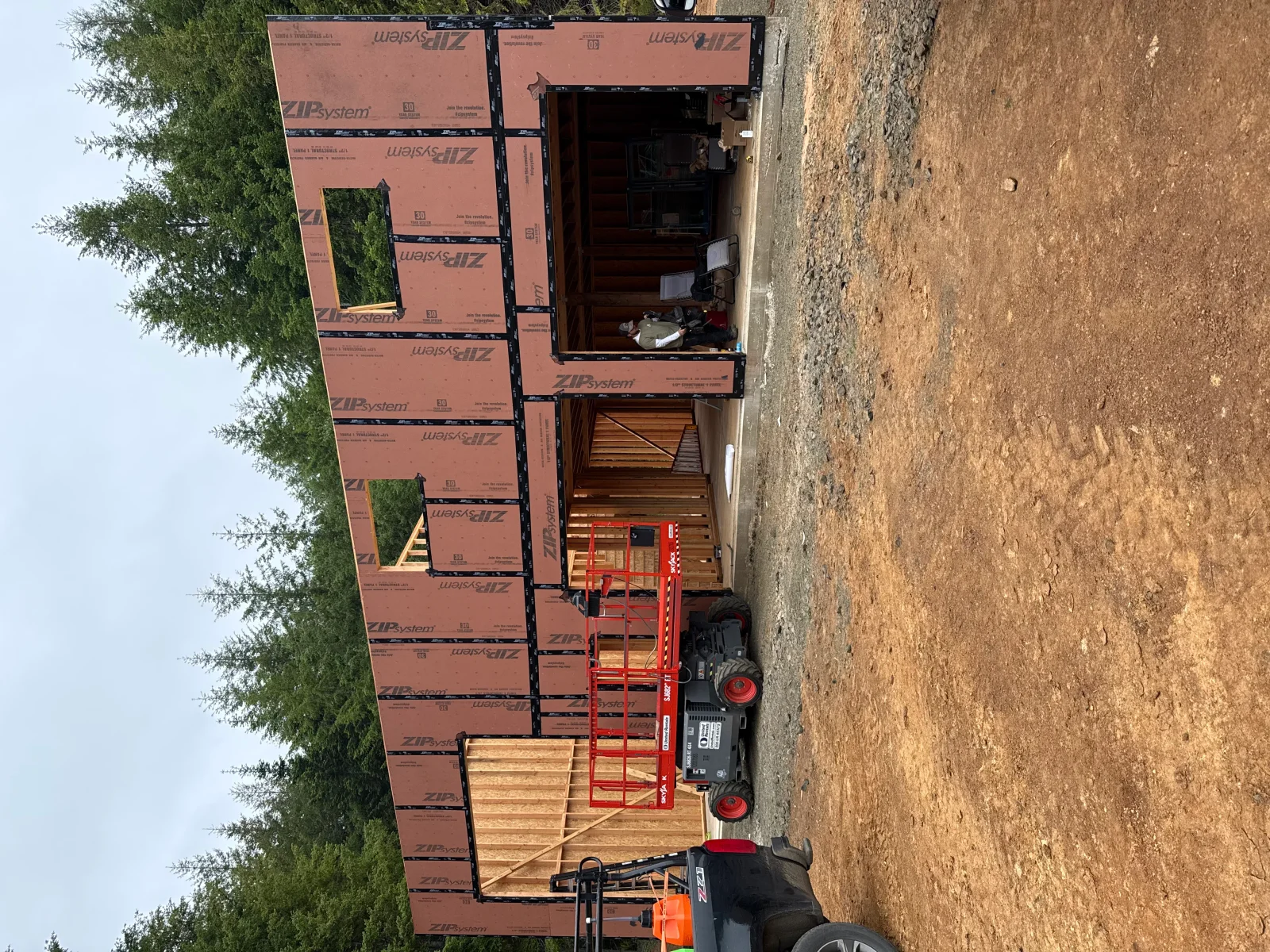 Wood-framed building with orange ZIP System sheathing and taped seams, rough window openings, and a red scissor lift on site
